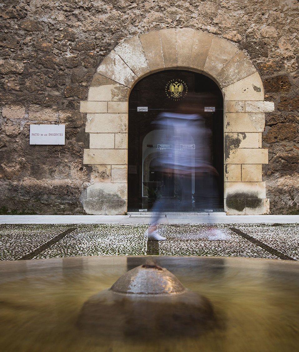 Patio del Hospital Real con fuente en el centro y puerta al fondo, a modo de ráfaga, una persona está pasando por la puerta